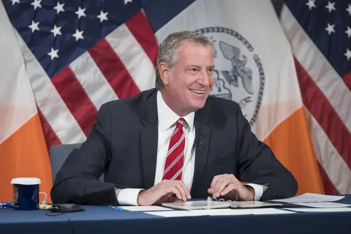 Mayor De Blasio, in a suit and tie, smiles while sitting at a dais during his daily press briefing